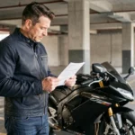 Motard en veste sombre consultant un document d'assurance moto dans un parking urbain en béton